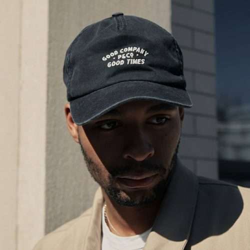 Model wearing washed black 5 panel hat with embroidered detail and leather strap closure.