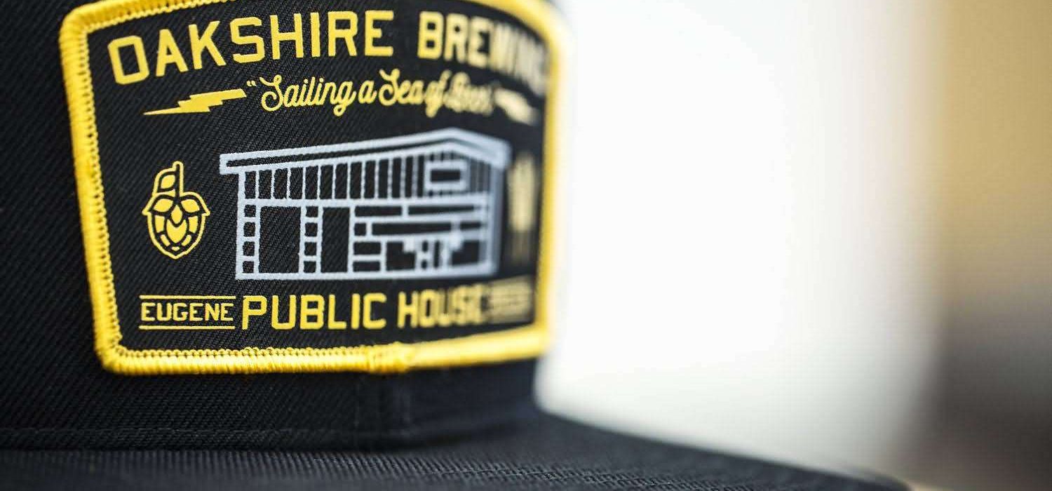 black-snapback-woven-patch-oakshire-brewing-public-house. Black structured snapback hat with detailed woven patch for Oakshire Brewing Public House, featuring yellow stitched border and brewery graphic