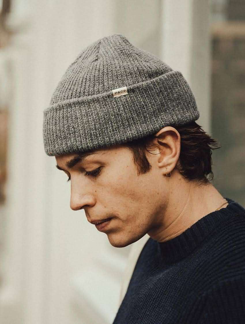 Heather grey ribbed knit beanie with small woven fabric label, worn by man in navy sweater, minimalist streetwear style