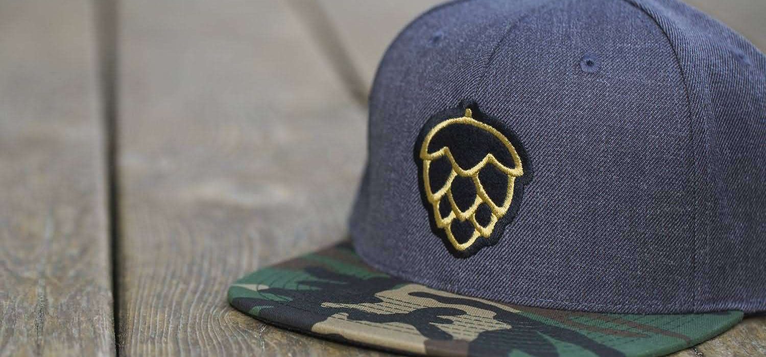 heather-navy-snapback-camo-brim-3d-embroidery-hop-logo. Heather navy snapback with camouflage brim and gold 3D puff embroidery hop cone logo