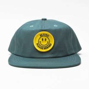 Symmetrical front view of a jade-colored unstructured 5-panel cap, featuring a front-centered circular yellow woven smiley face patch with black text reading 'Be Kind' and 'Please Rewind'. The flat visor with multi-row stitching is fully presented.