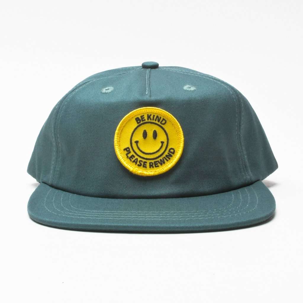 Symmetrical front view of a jade-colored unstructured 5-panel cap, featuring a front-centered circular yellow woven smiley face patch with black text reading 'Be Kind' and 'Please Rewind'. The flat visor with multi-row stitching is fully presented.
