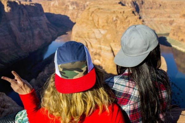 Two individuals overlooking a canyon, wearing a custom 5-panel camo mesh trucker hat and a plain grey twill flat brim cap.