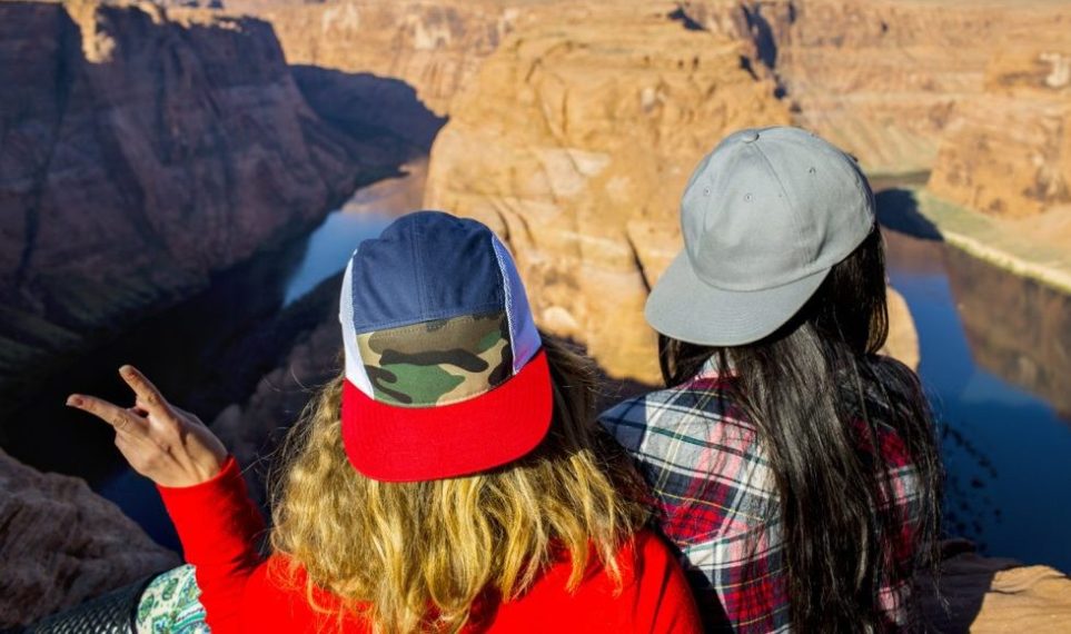 Two individuals overlooking a canyon, wearing a custom 5-panel camo mesh trucker hat and a plain grey twill flat brim cap.