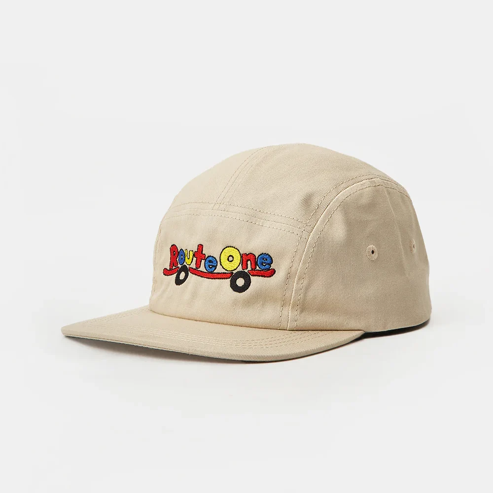 Angled front view of a beige cotton twill 5-panel camp cap featuring a flat brim and colorful "Route One" skateboard flat embroidery on the front panel.