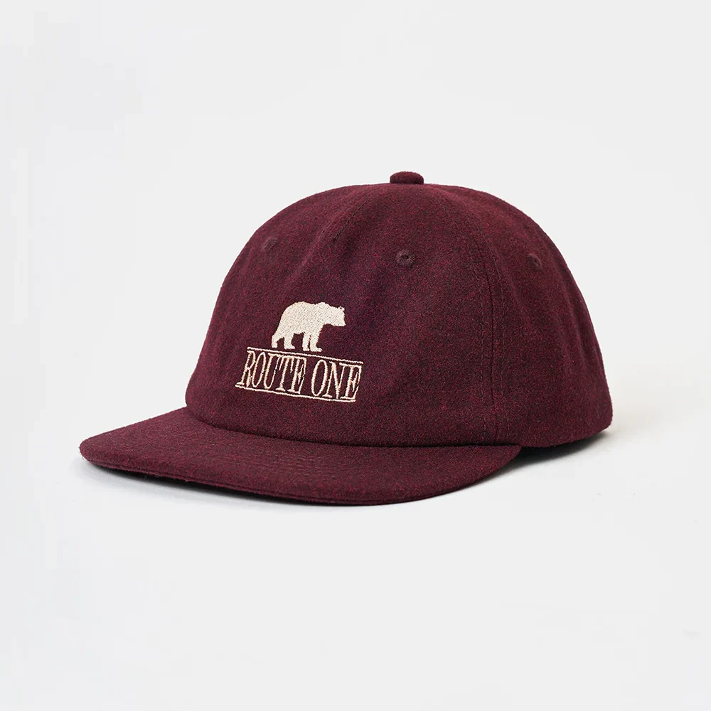 Angled front view of an unstructured burgundy wool melton 6-panel dad hat featuring a flat brim and crisp high-density beige "Route One" bear embroidery.