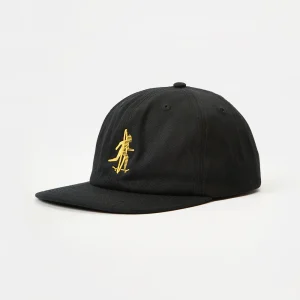 Front three-quarter view of a black unstructured cotton twill flat brim cap featuring gold flat embroidery of a surfer-skater figure on the front panel, with visible low-profile crown shape and flat visor angle.